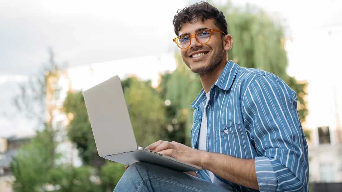 Junger Mann mit Laptop sitzt draussen und arbeitet am Laptop