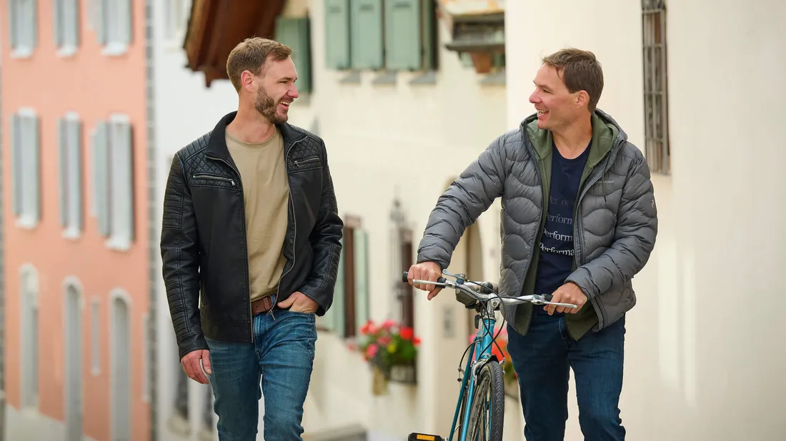 Zwei Männer spazieren durch die Stadt, einer schiebt ein Fahrrad