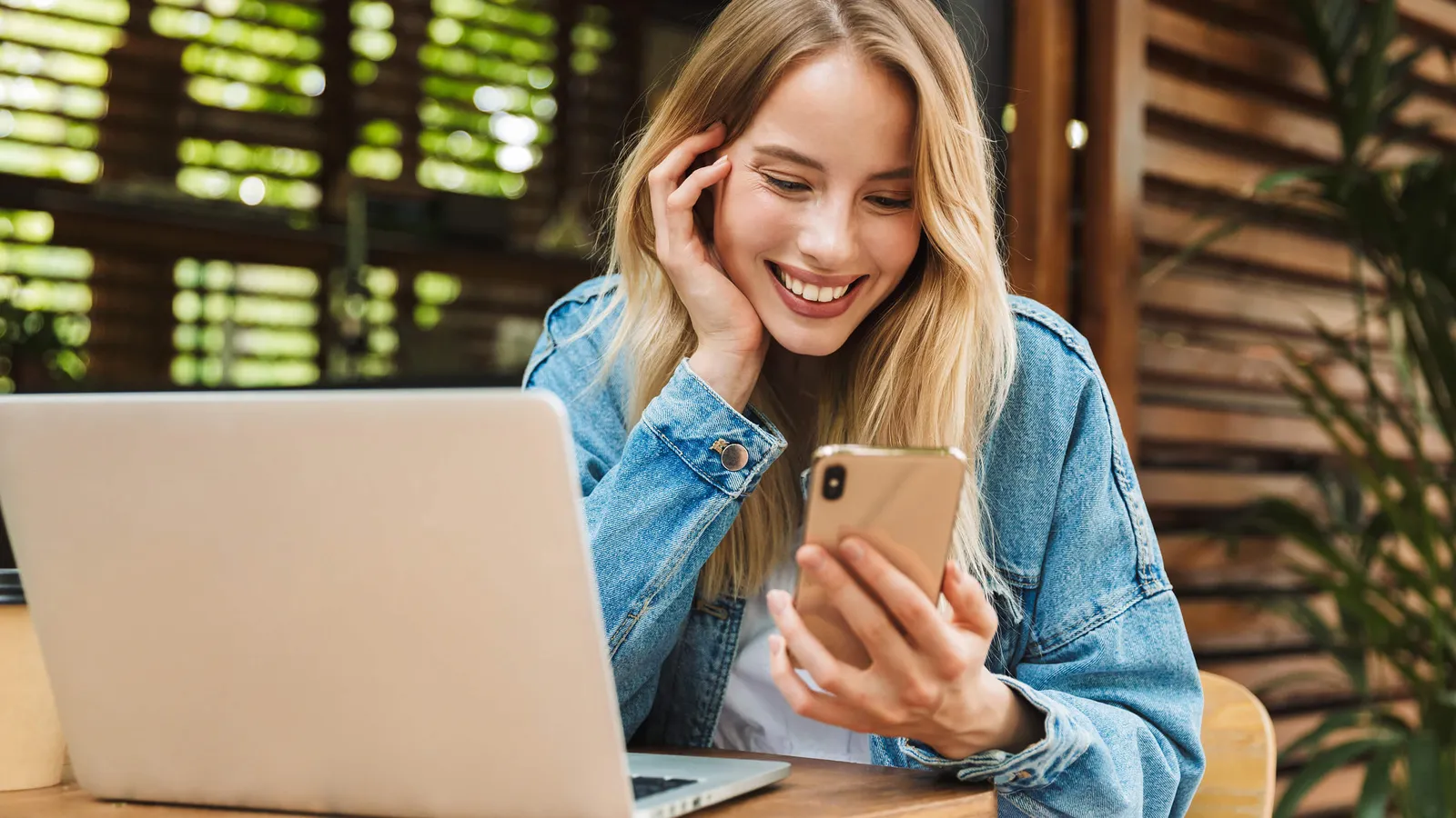 Junge Frau sitzt an einem Tisch mit Laptop und blickt auf ihr Handy