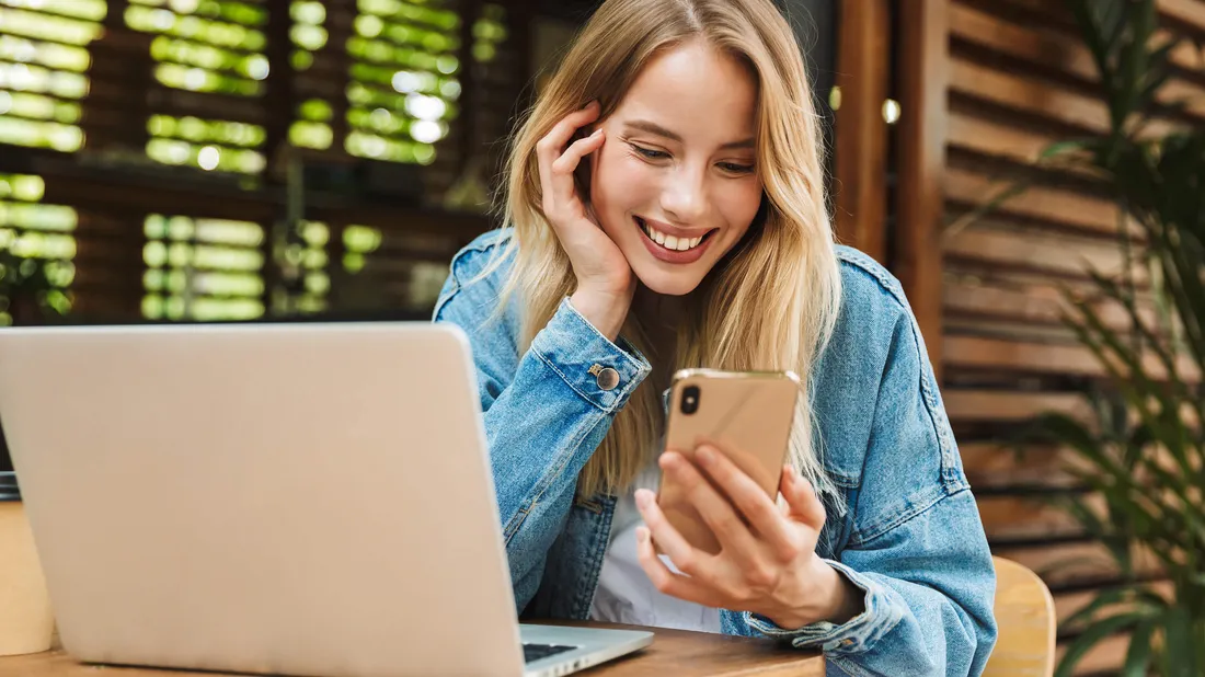 Junge Frau sitzt an einem Tisch mit Laptop und blickt auf ihr Handy