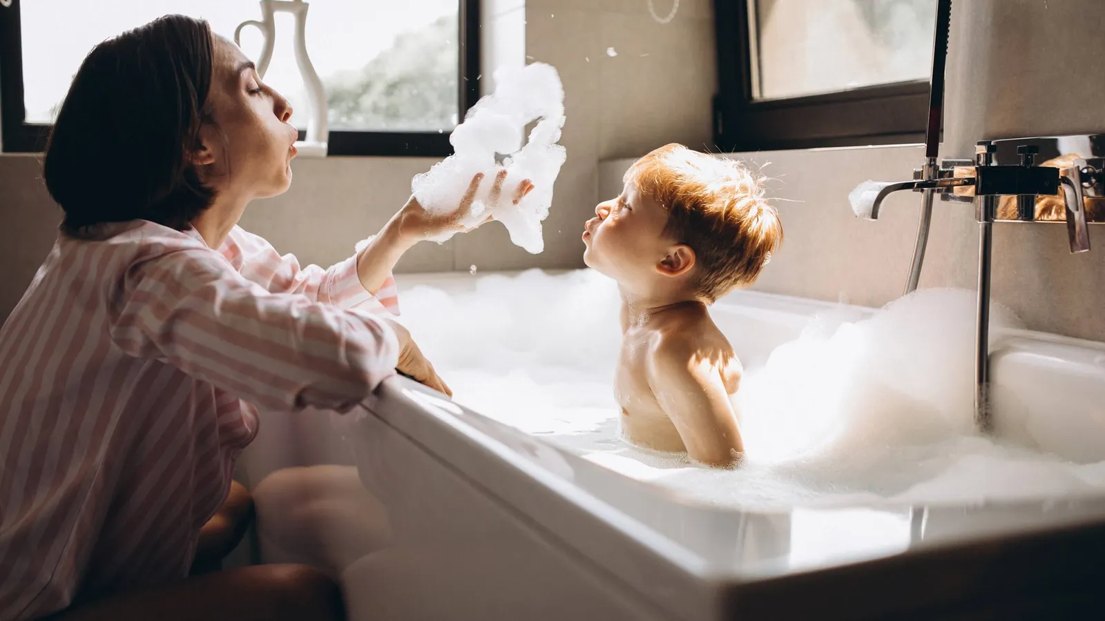 Ein Kind in der Badewanne mit viel Schaum, die Mutter bläst in den Schaum